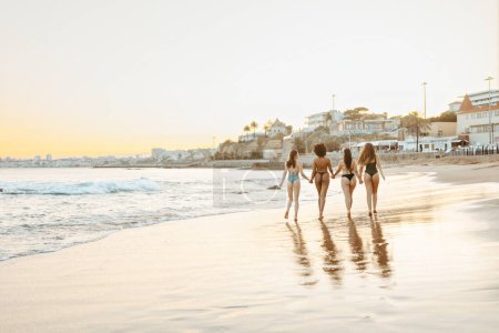 Photo for Back view of four young women friends walking, holding hands and enjoying summer time on the beach at sunset, full length shot, free copy space, banner - Royalty Free Image