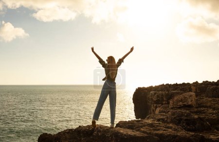 Photo for Freedom Concept. Young woman standing on the edge of rock ledge at sunset and looking at ocean, unrecognizable blonde female traveller raising hands up, enjoying outdoor adventures, copy space - Royalty Free Image