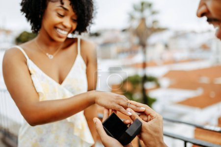 Foto de Feliz joven afroamericano chico poner anillo en el dedo de la señora sorprendida, propone matrimonio en la ciudad, disfruta de la fecha, al aire libre. Di que sí, amor, relaciones, vacaciones y romance, compromiso - Imagen libre de derechos