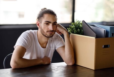 Photo for Dismissal, Economic crisis. Unhappy hispanic young man sitting upset near personal belongings in box after being fired at office interior. Unemployed guy feeling depressed after losing his job - Royalty Free Image