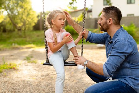 Photo for Playground injury. Father talking with daughter sitting on swing, girl touching injured knee and looking at dad. Child running and fell while playing in park - Royalty Free Image