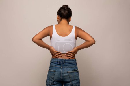 Téléchargez les photos : Vue arrière de la jeune femme noire massant son bas du dos, souffrant de tension musculaire ou de scoliose, isolée sur fond de studio gris. Arthrite, problèmes structurels et lésions discales - en image libre de droit