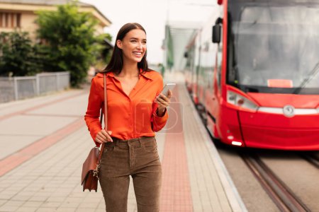 Photo for Happy Young Passenger Lady With Cellphone Booking Ticket For Tram Ride Via Application Standing Near Public Transport Stop Outdoor, Looking Aside. Easy Transportation And City Lifestyle Concept - Royalty Free Image
