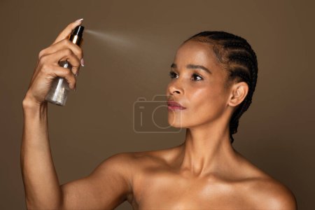 Téléchargez les photos : Belle femme afro-américaine d'âge moyen utilisant un spray à l'eau thermale, hydratant sa peau, debout sur fond brun. Concept beauté et soin de la peau - en image libre de droit