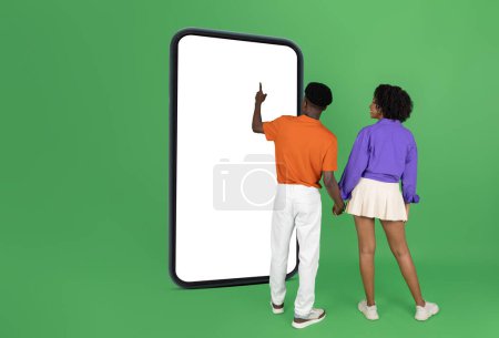 Foto de Millennial afroamericano chico y la señora en los dedos de punta casual en el teléfono inteligente grande con pantalla vacía, aislado en fondo de estudio verde, longitud completa. App, blog, sitio web en venta, redes sociales - Imagen libre de derechos