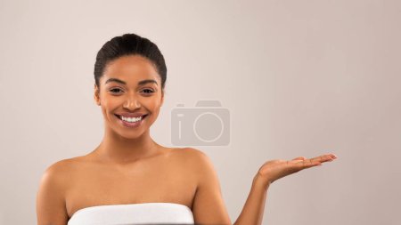 Téléchargez les photos : Positif joyeux attrayant jeune femme noire enveloppé dans une serviette blanche montrant quelque chose sur sa main de la paume et souriant à la caméra, recommandant de beaux produits de beauté, isolé sur gris, espace de copie - en image libre de droit