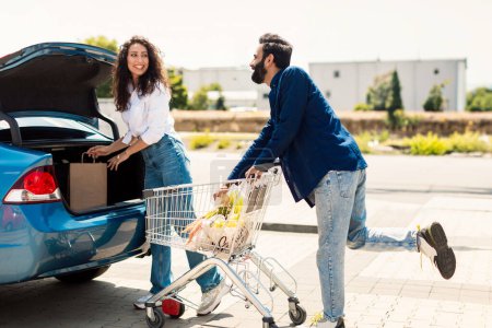 Téléchargez les photos : Jeune couple élégant du Moyen-Orient avec panier plein de nourriture fraîche, emballage des produits dans la voiture sur le parking extérieur, rentrer à la maison après des achats réussis - en image libre de droit