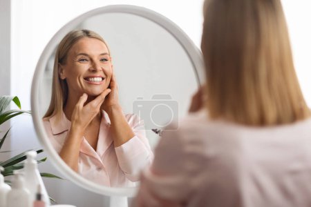 Foto de Hermosa mujer de mediana edad que mira en el espejo en su suave piel lisa, atractiva cara femenina madura conmovedora y sonriendo a la reflexión, disfrutando de la rutina de cuidado de la piel en el hogar, enfoque selectivo - Imagen libre de derechos