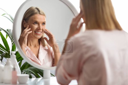 Foto de Rutina de autocuidado. Atractiva mujer madura mirando en el espejo en casa, feliz edad media hembra que inspecciona la cara, tocando su suave piel lisa y sonriente, disfrutando de tratamientos para el cuidado de la piel Resultado - Imagen libre de derechos
