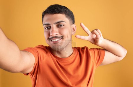 Téléchargez les photos : Jeune homme brésilien insouciant prenant selfie, montrant geste de paix et souriant, ayant une humeur ludique, gars positif posant sur fond de studio orange - en image libre de droit