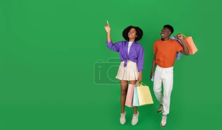 Photo for Cheerful millennial black couple shopaholics with packages with purchases point finger on empty space, jumping, freeze in air, isolated on green studio background. Shopping together, emotions, sale - Royalty Free Image