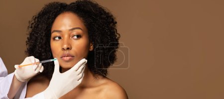 Photo for Hot pretty young black woman get lip filler injection, beautitian hands holding syringe next to patient mouth, brown background, web-banner for aesthetic cosmetology, copy space - Royalty Free Image
