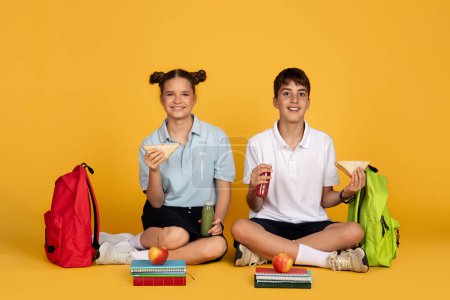 Photo for Happy teen european boy and girl with backpacks eating apple and sandwich, drink juice bottle, isolated on yellow studio background. Friendship and lunch, education at school, break for food - Royalty Free Image