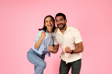 Photo for Thrilled happy beautiful loving millennial couple celebrating success together isolated on pink studio background. Emotional lucky handsome guy and pretty woman clenching fists and exclaiming - Royalty Free Image