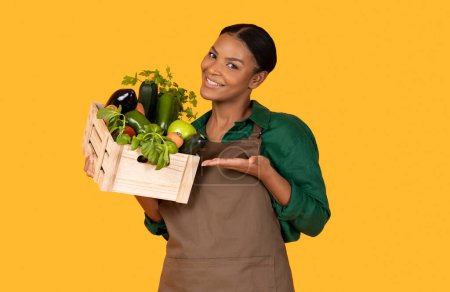 Téléchargez les photos : La nourriture biologique. Femme agricultrice afro-américaine souriante montrant sa récolte de jardin gestuelle à la boîte avec des légumes et des fruits frais sur fond jaune studio. Jardinage et agriculture Affaires - en image libre de droit