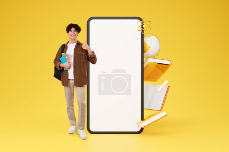 Photo for College Student Guy Posing With Books And Huge Smartphone Gesturing Thumbs Up, Approving Application, Empty Screen For Mockup Standing On Yellow Studio Background With Educational Icons. Collage - Royalty Free Image