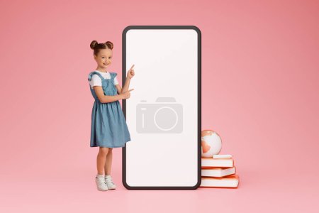 Photo for Online Study. Cute schoolgirl pointing at large smartphone touchscreen on pink background showing blank screen mockup for educational application advert. Collage with books and world globe icon, - Royalty Free Image