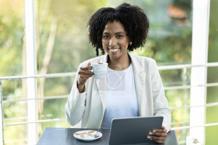 Téléchargez les photos : Portrait d'une belle femme d'affaires afro-américaine millénaire en tenue formelle blanche assise à table, buvant du café au café, utilisant une tablette numérique, travaillant en ligne, espace de copie - en image libre de droit