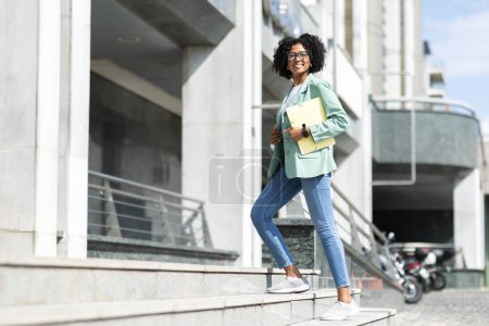 Téléchargez les photos : Positif joyeux attrayant jeune femme noire portant une tenue décontractée élégante et des lunettes avec des cahiers dans sa main employé allant au bureau le matin, espace de copie. Travail, carrière - en image libre de droit