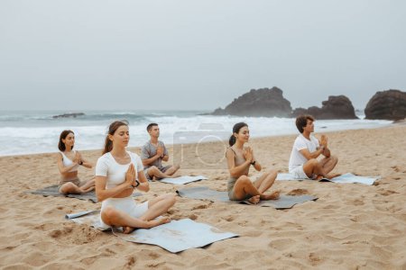 Foto de Hombres y mujeres jóvenes pacíficos meditando juntos en la playa, practicando yoga en posición de loto con los ojos cerrados, sentados sobre esteras y ejercitando diferentes asanas, espacio libre - Imagen libre de derechos