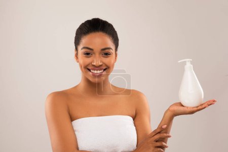 Téléchargez les photos : Attrayant souriant souriant millénaire femme noire enveloppé serviette blanche tenant bouteille avec des produits de beauté, afro-américaine dame appréciant la peau du corps soyeux après nouvelle lotion, isolé sur fond gris - en image libre de droit