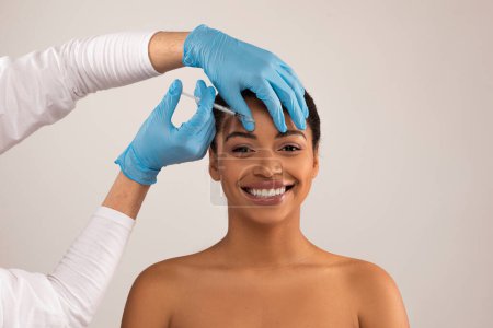 Photo for Young black woman getting beauty cosmetic injection in forehead. Cheerful attractive millennial half-naked lady have filler in interbrow area at clinic, grey background. Beauty treatments concept - Royalty Free Image