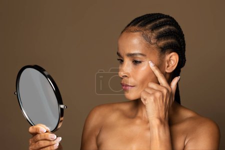 Téléchargez les photos : Vieillissement cutané. Belle femme noire d'âge moyen tenant miroir et regardant ses rides près des yeux, vérifiant le visage et remarquant des changements d'âge, debout sur fond brun - en image libre de droit