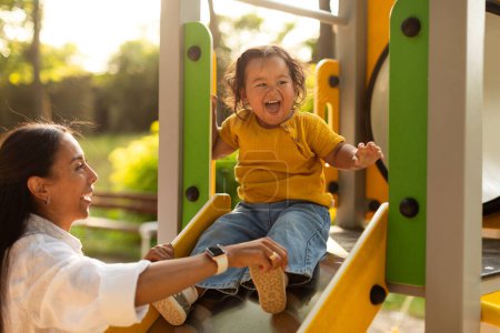 Photo for Japanese Mother Riding Happy Baby Daughter Girl Down The Slide Spending Time Together At Playground Outside. Mommy Or Babysitter Woman Playing And Bonding With Adorable Toddler Infant - Royalty Free Image
