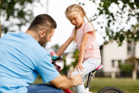 Téléchargez les photos : Petite fille assise à vélo ressentant une douleur à la jambe, est tombée alors qu'elle roulait à vélo dans le parc, père inquiet réconfortant sa fille, touchant et vérifiant son genou - en image libre de droit
