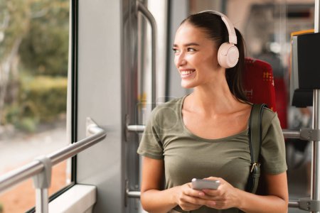 Téléchargez les photos : Heureux Passager. Lady assis sur le siège dans le tramway moderne portant des écouteurs sans fil et écoutant de la musique en ligne par téléphone, se rendre au travail. Concept de transport urbain confortable - en image libre de droit