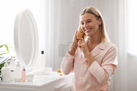 Photo for Smiling beautiful middle aged woman combing her smooth hair with bamboo brush at home, happy mature blonde woman wearing sil pajamas using comb while sitting at dressing table in bedroom - Royalty Free Image