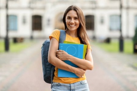 Beautiful Young Female Student With Backpack And Workbooks Posing Outdoors Near College Building, Happy Millennial Woman Smiling To Camera, Enjoying Educational Programs And Studentship, Copy Space