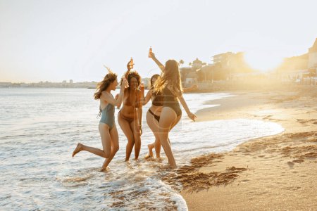 Foto de Jovencitas alegres multirraciales en trajes de baño bailando, sosteniendo botellas de cerveza, disfrutando de la fiesta de verano en la playa, amigas encontrándose y divirtiéndose en la costa, espacio libre - Imagen libre de derechos