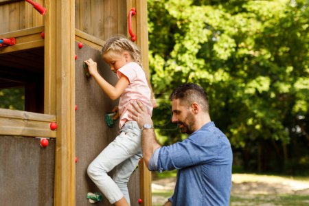 Photo for Father and daughter walking in park, girl trying to climb on wall at playground and dad playing, family spending time together outdoors. Hand and eye coordination, motor skills development - Royalty Free Image
