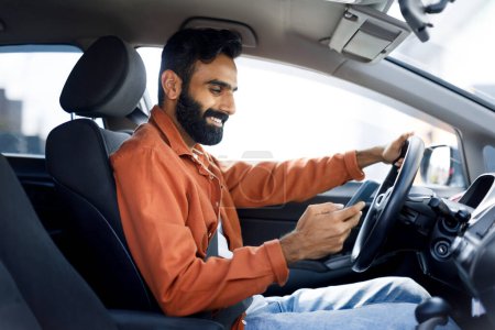 Téléchargez les photos : Sourire gars indien en utilisant la messagerie smartphone et de vérifier les e-mails tout en conduisant une voiture. Vue latérale du conducteur du Moyen-Orient homme regardant son téléphone portable pour la navigation, la recherche meilleure façon - en image libre de droit