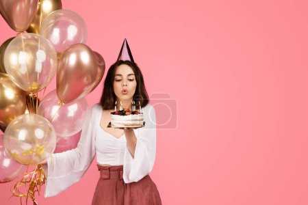 Photo for Cheerful millennial caucasian brunette woman in hat with many balloons blows out candles on cake, isolated on pink background, studio. Ad and offer, celebration holiday, birthday party - Royalty Free Image