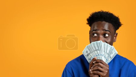 Photo for Lottery Luck. Happy young african american man has just won the lottery jackpot, holding bunch of cash dollar banknotes over his face, looking at copy space, isolated on yellow background, banner - Royalty Free Image