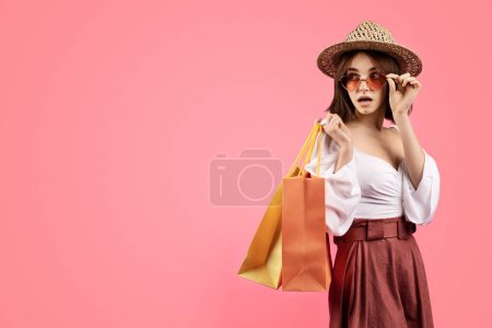 Photo for Cheerful millennial caucasian woman with open mouth in hat with many packages takes off glasses, looks at empty space, isolated on pink background, studio. Ad and offer, lifestyle, shopping, sale - Royalty Free Image