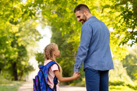 Foto de Feliz padre de mediana edad y colegiala hija caminando a la escuela, tomados de la mano y mirándose entre sí. Concepto de paternidad y vínculo. Vías de aprendizaje - Imagen libre de derechos