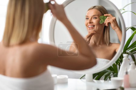 Téléchargez les photos : Souriant visage de massage féminin d'âge moyen avec grattoir en pierre de quartz gua sha pour les soins de la peau, heureuse femme mûre regardant dans le miroir, faire des soins de beauté à la maison, profiter d'une peau saine et éclatante - en image libre de droit