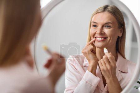 Téléchargez les photos : Belle femme mûre regardant le miroir et appliquant le baume à lèvres hydratant, souriant attrayant moyen âge Lady tenant Chapstick, en utilisant le rouge à lèvres hygiénique tout en faisant la routine de beauté domestique - en image libre de droit