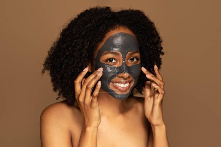 Foto de Sonriente positiva atractiva milenaria topless dama afroamericana utilizando máscara negra, aislado sobre fondo marrón. Mascarilla facial despegable para una limpieza profunda de la piel, concepto de cuidado facial - Imagen libre de derechos