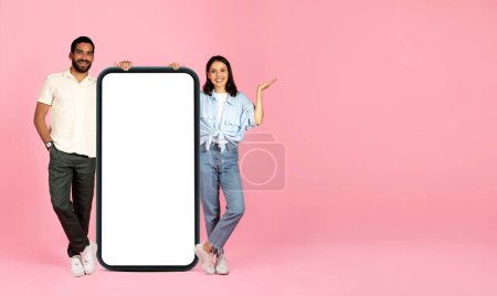 Photo for Positive cute beautiful millennial couple posing with huge cell phone with white blank screen, pointing at copy space for advertisement, isolated on pink studio background, panorama, mockup - Royalty Free Image