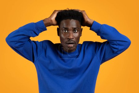 Photo for Shocked appalled handsome young black guy wearing casual blue sweater touching his head and grimacing, isolated on yellow studio background, african american man hear devastating news - Royalty Free Image