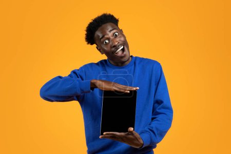 Photo for Crazy funny handsome young african american guy wearing blue sweater showing modern pc digital tablet with empty black screen and grimacing, showing nice app, yellow background - Royalty Free Image