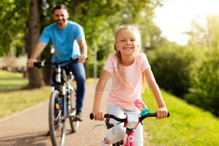 Photo for Day in the park. Happy family of caucasian father and girl child riding bicycles outdoors in the summer, smiling at camera. Dad and daughters park adventure - Royalty Free Image