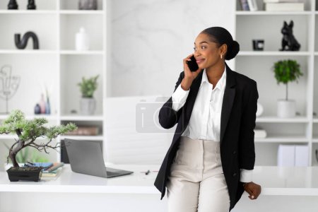 Photo for Business communication concept. Happy black businesswoman talking on cellphone standing near her workplace in light office, looking aside, copy space. Entrepreneur communicating for work - Royalty Free Image