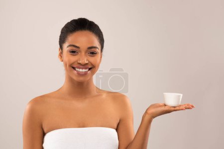 Photo for Smiling cheerful attractive young black woman wrapped in towel holding white jar with beauty product, using organic moisturizing face cream cosmetics for youthful looking skin, studio background - Royalty Free Image