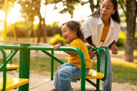 Photo for Babyhood Joy And Fun. Happy Adorable Asian Baby Girl Having Fun Riding Sitting On Carousel, Mother Pushing Merry-Go-Round At Outdoor Playground. Selective Focus On Laughing Child - Royalty Free Image