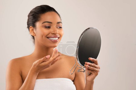 Foto de Atractiva joven negra sonriente envuelta en toalla de baño aplicando crema facial, mirando al espejo y sonriendo. Mujer afroamericana semidesnuda haciendo rutina de cuidado de la piel, fondo gris, espacio para copiar - Imagen libre de derechos